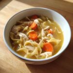 A close-up of a white bowl filled with rich broth, shredded chicken, egg noodles, and bright orange carrots from the slow cooker chicken noodle soup.