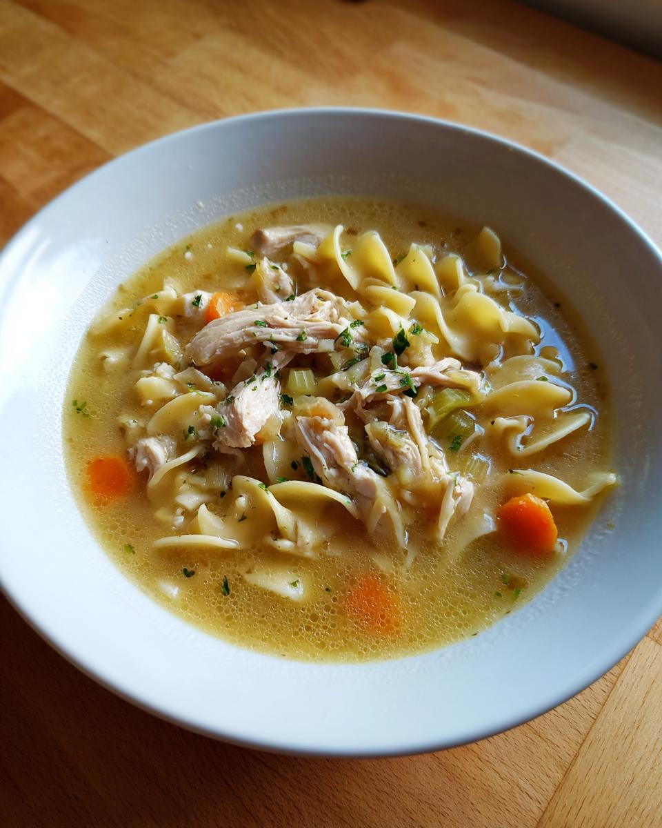 A close-up of a white bowl filled with rich broth, shredded chicken, egg noodles, and carrots from the slow cooker chicken noodle soup.