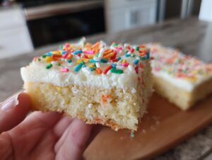 Close-up of a soft batch sugar cookie bars slice held by a hand, topped with white frosting and colorful sprinkles.
