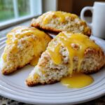 Close-up of three soft lemon scones stacked on a white plate, generously drizzled with bright yellow lemon glaze.