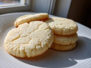 A stack of soft, pale yellow sugar cookie cutouts resting on a white plate near a window.