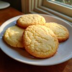 Close-up of four soft, golden sugar cookie rounds sprinkled with sugar on a white plate.