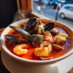 A steaming white bowl filled with vibrant red seafood soup featuring mussels, shrimp, clams, and chunks of fish.