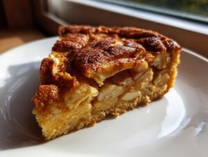 A close-up slice of moist Swedish apple cake, featuring visible apple chunks and a cinnamon-dusted top, served on a white plate.