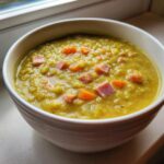 Close-up of a thick, yellow-green pea soup topped with diced ham and bright orange carrots in a white bowl.