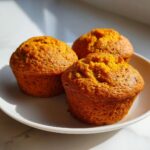 Close-up of three freshly baked, golden-orange sweet potato muffins resting on a white plate.