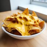 A white bowl overflowing with tortilla chips generously smothered in bright yellow nacho cheese sauce.