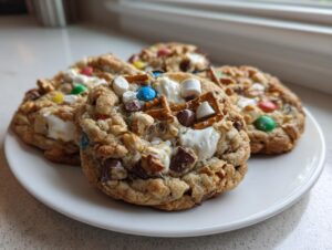 Four ultimate kitchen sink cookies loaded with M&Ms, marshmallows, pretzels, and chocolate chunks on a white plate.