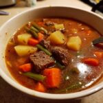 Close-up of a hearty bowl of vegetable beef soup featuring chunks of beef, potatoes, carrots, and green beans in a rich broth.