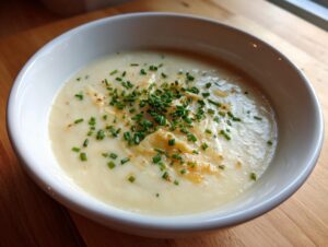 A close-up of a bowl of velvety cauliflower soup garnished with shredded cheese and fresh chopped chives.