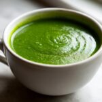 Close-up of vibrant, creamy green spinach soup served in a white mug, showing a slight swirl on top.
