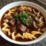 Close-up of a white bowl filled with rich, dark broth, chunks of tender beef, and egg noodles, garnished with fresh parsley for the beef noodle soup.