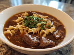 Close-up of a white bowl filled with rich, dark broth, tender chunks of beef, egg noodles, and fresh parsley, showcasing the beef noodle soup.