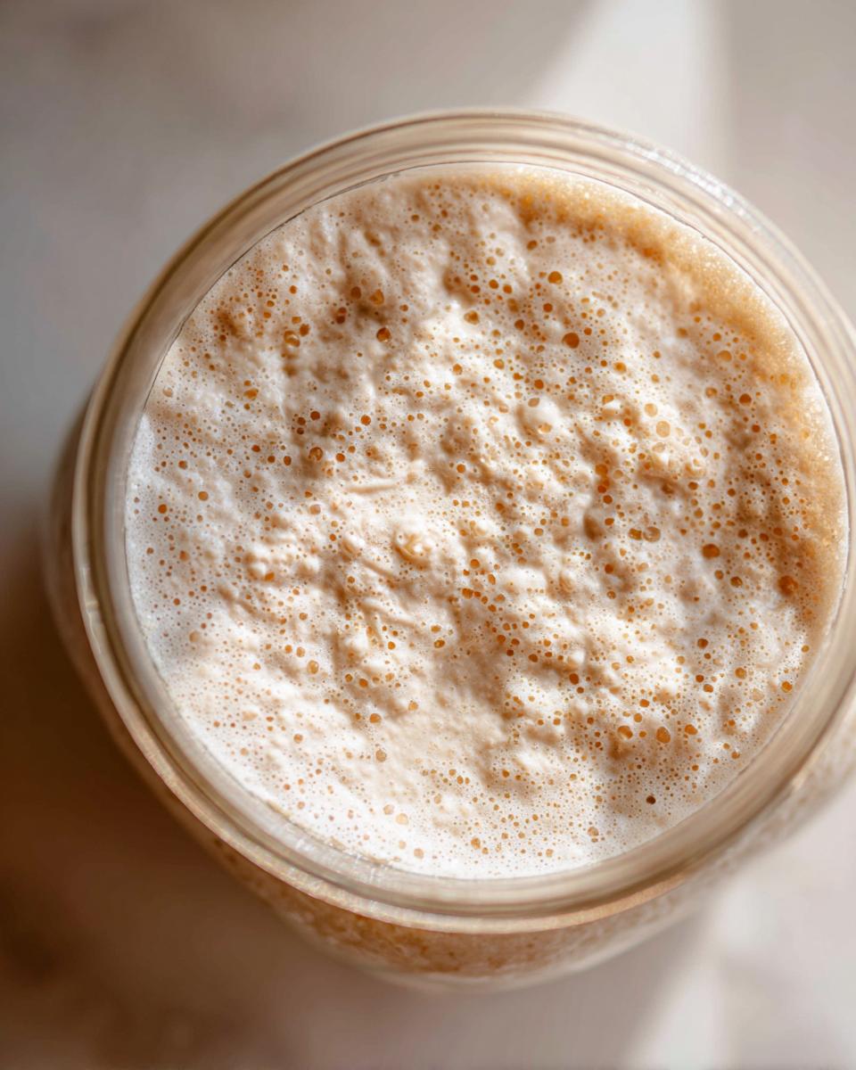 Overhead view of a highly active sourdough starter in a glass jar, showing a thick layer of foam and many small bubbles.
