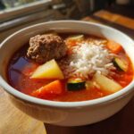 Close-up of a bowl of rich, red albondigas soup featuring a large meatball, white rice, potatoes, carrots, and zucchini.