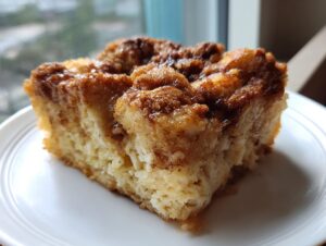 A close-up of a square slice of baked french toast casserole with a gooey, dark cinnamon-sugar topping on a white plate.