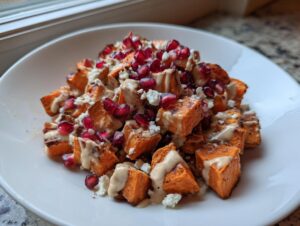 Close-up of roasted cubes in a sweet potato salad topped with pomegranate seeds, feta, and a creamy dressing.