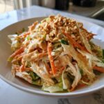 A vibrant bowl of thai chicken salad featuring shredded chicken, carrots, cabbage, and a creamy peanut dressing, topped with crushed peanuts.