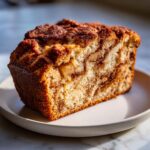 A close-up slice of moist apple bread featuring a cinnamon swirl and visible chunks of apple, sitting on a light plate.