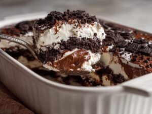 A spoonful of rich Baileys Chocolate Cheesecake Trifle being lifted from a white baking dish, showing layers of cream, chocolate pudding, and cookie crumbs.