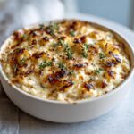 Close-up of a baked caramelized onion dip in a white bowl, featuring a bubbly, browned cheese crust and fresh thyme garnish.
