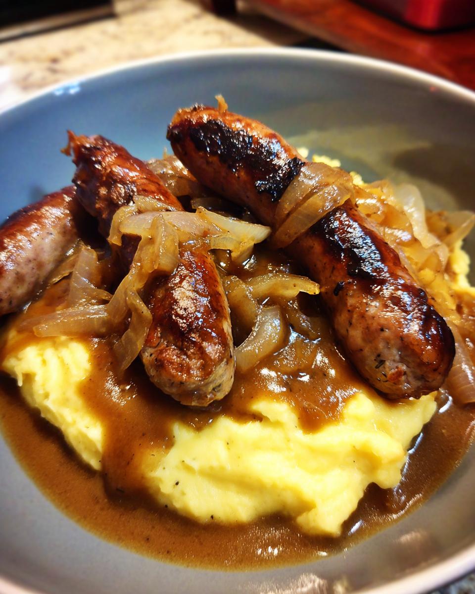 Close-up of a bowl of bangers and mash featuring browned sausages, creamy mashed potatoes, and onion gravy.