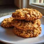 A stack of three golden brown, chewy oatmeal cookies resting on a white plate, highlighted by natural window light.