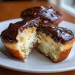 A Boston cream pie cupcake cut in half showing the vanilla cake, pastry cream filling, and chocolate glaze topping.