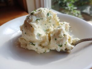 A close-up of creamy boursin mashed potatoes mixed with green herbs, served on a white plate with a spoon.