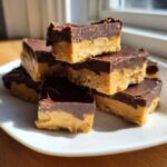 A stack of rich, layered buckeye fudge squares featuring a peanut butter base and chocolate topping on a white plate.