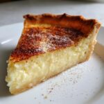 Close-up of a creamy slice of buttermilk pie on a white plate, dusted with powdered sugar.