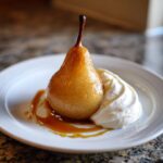 A beautifully caramelized poached pear served on a white plate with a dollop of whipped cream and caramel sauce.