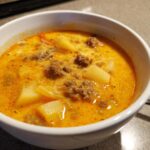 Close-up of a white bowl filled with creamy, orange-hued cheeseburger soup containing ground beef and chunks of potato.