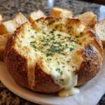 Close-up of a hot, cheesy bread dip served inside a hollowed-out sourdough loaf, garnished with parsley, with bread pieces surrounding it.