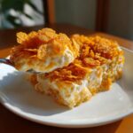 A spoonful of creamy cheesy potato casserole topped with crispy cornflakes being lifted from a serving on a white plate.