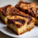 Four rich, chewy snickerdoodle blondies squares with a prominent cinnamon swirl topping on a white plate.