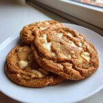 Close-up of three golden brown white chocolate macadamia nut cookies stacked slightly on a white plate.