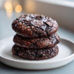 A stack of three rich, dark chocolate brownie cookies sprinkled with flaky sea salt, sitting on a white plate.
