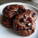 Close-up of rich, dark chocolate cherry cookies with melted chocolate chips and dried cherries, one cookie broken open.