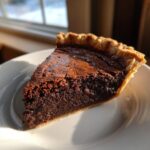 A close-up slice of rich, dark chocolate chess pie with a flaky crust, sitting on a white plate bathed in sunlight.