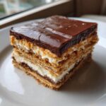 A close-up of a square slice of chocolate eclair cake showing distinct layers of graham crackers, cream filling, and a thick chocolate glaze.