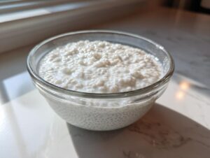 A clear glass bowl filled with creamy white coconut chia pudding, set near a window.