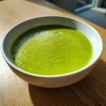 Close-up of a white bowl filled with bright green, creamy asparagus soup on a wooden table.