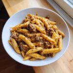 A close-up shot of a white bowl filled with creamy beef pasta, featuring penne noodles coated in a rich sauce.