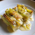 A close-up of tender braised leeks baked with a creamy, cheesy sauce, served on a white plate.