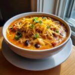 A close-up of a bowl of creamy buffalo chicken chili topped with shredded cheddar cheese and sliced green onions.