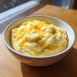 A white bowl filled with creamy, fluffy cheese grits topped with melted cheddar cheese.