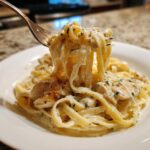 A fork lifting a generous portion of creamy chicken alfredo pasta, showing fettuccine noodles and sliced chicken.