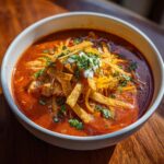 Close-up of a bowl of rich, red creamy chicken enchilada soup topped with tortilla strips, cheese, sour cream, and cilantro.