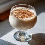 A glass filled with creamy, light-colored coquito, topped generously with ground cinnamon.